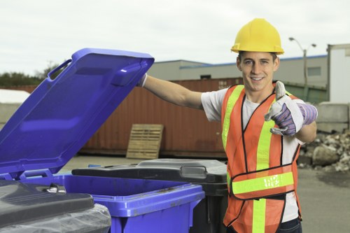 A young male waste management worker wearing a yellow safety helmet, a high-visibility orange vest with reflective strips, and purple work gloves, is smiling and giving a thumbs-up while standing outdoors on a paved surface. He is positioned next to opened blue and black rubbish bins, which appear to be made of durable plastic with textured surfaces. In the background, there are industrial buildings, a partially visible fence, and some construction materials, indicating an area designated for waste collection or clearance. The scene suggests a professional approach to rubbish removal, consistent with services offered by Commercial Waste Borehamwood in the local area. The lighting is natural, with an overcast sky providing diffuse illumination, ensuring clear visibility of the objects and the worker. The overall setting emphasizes a tidy, organized environment suitable for commercial waste disposal activities, highlighting the company's local expertise in rubbish removal services around Borehamwood and the wider Hertfordshire region.
