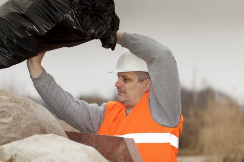 A male worker wearing a white safety helmet and an orange high-visibility vest is lifting a black plastic rubbish bag, which appears to be filled with waste materials, from a pile of mixed refuse. The waste pile contains various materials, including a beige fabric sack and other unidentifiable debris, situated outdoors on a concrete or paved surface. The background is blurred, showing a cloudy sky and an open landscape with dry grass, indicating a rural or industrial environment in Borehamwood. The worker's focused expression reflects careful handling of waste removal tasks, consistent with professional rubbish collection services by Commercial Waste Borehamwood. The scene emphasizes waste management activity, highlighting the collection and disposal of rubbish in a straightforward, factual manner suitable for a commercial waste removal context.