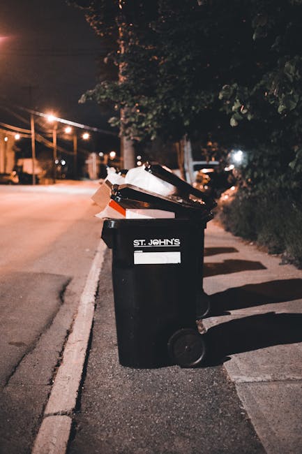 A black wheeled rubbish bin with a white label and the words 'ST. JOHN'S' printed on the front is positioned on a paved sidewalk next to a curb in a residential area at night. The bin is filled with a